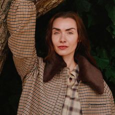 A woman wearing a jacket and tartan shirt from Whistles while holding onto a branch of a tree above her