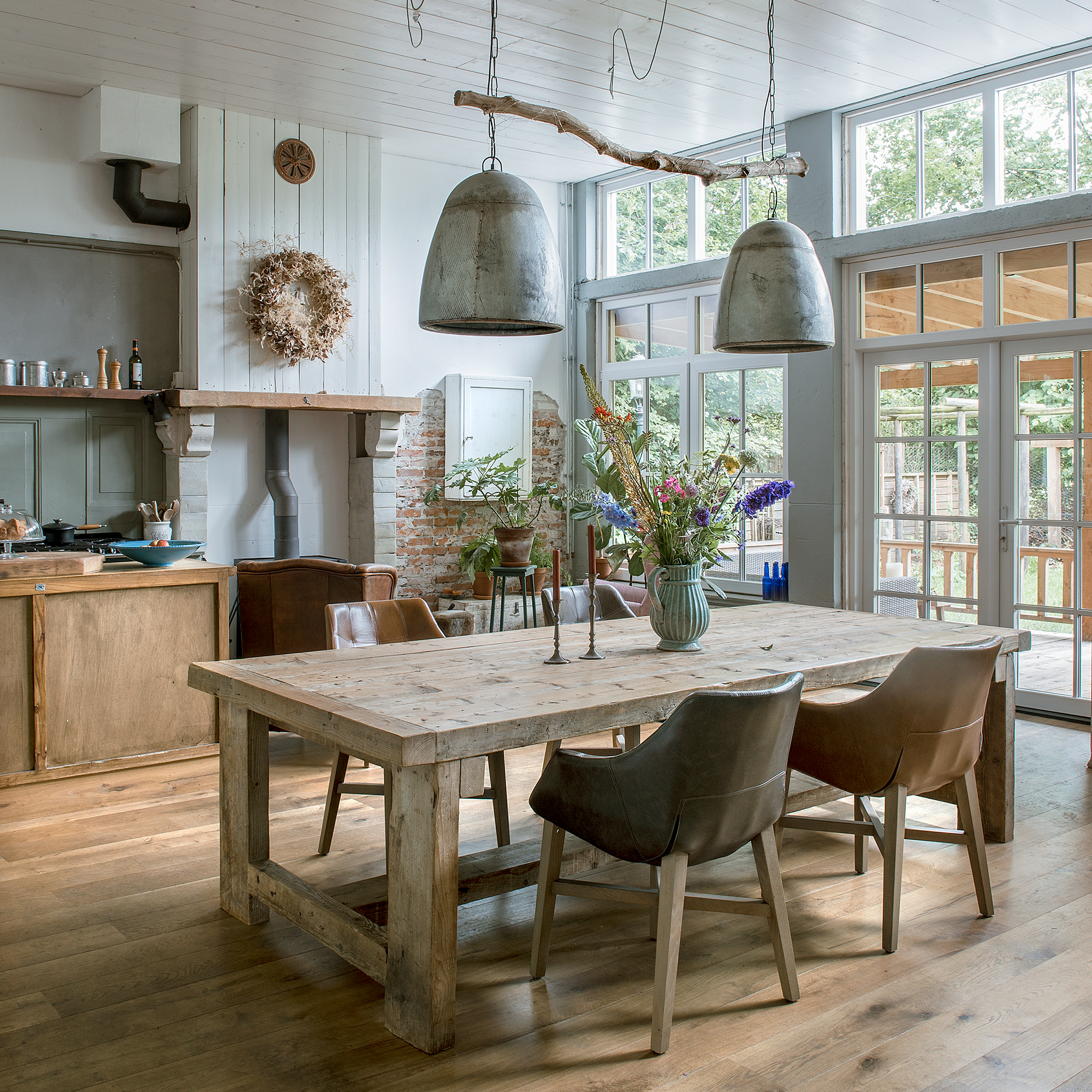 Open plan dining area with large wooden table, metal industrial pendants above, floor to ceiling windows and wooden floor