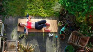 Woman holding two dumbbells doing a chest press lying down on yoga mat in back garden