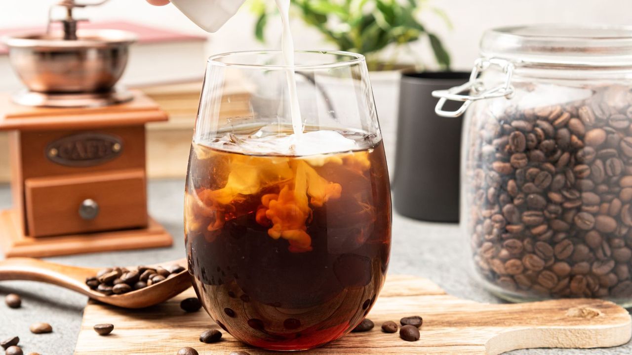 An example of how to make cold brew coffee, milk is being poured into a glass of cold brew coffee with coffee beans and measuring instruments behind it