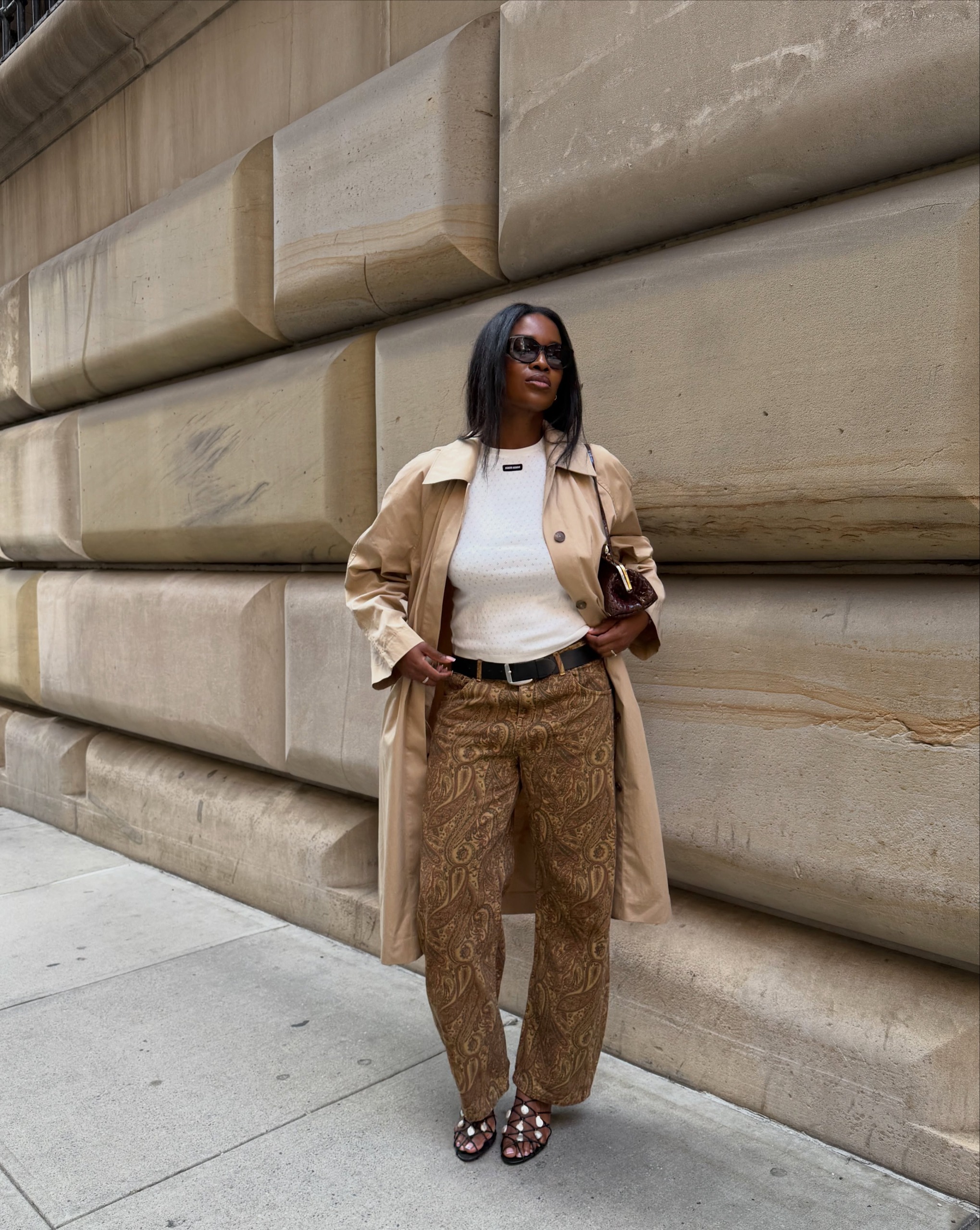 An image of an influencer wearing brown paisley trousers with a white T-shirt, beige trench coat and black heeled sandals
