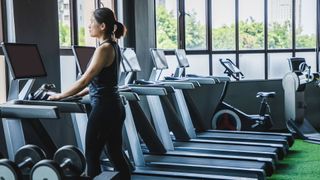 woman walking on treadmill in the gym, surrounded by weights and stationary bikes in front of large window