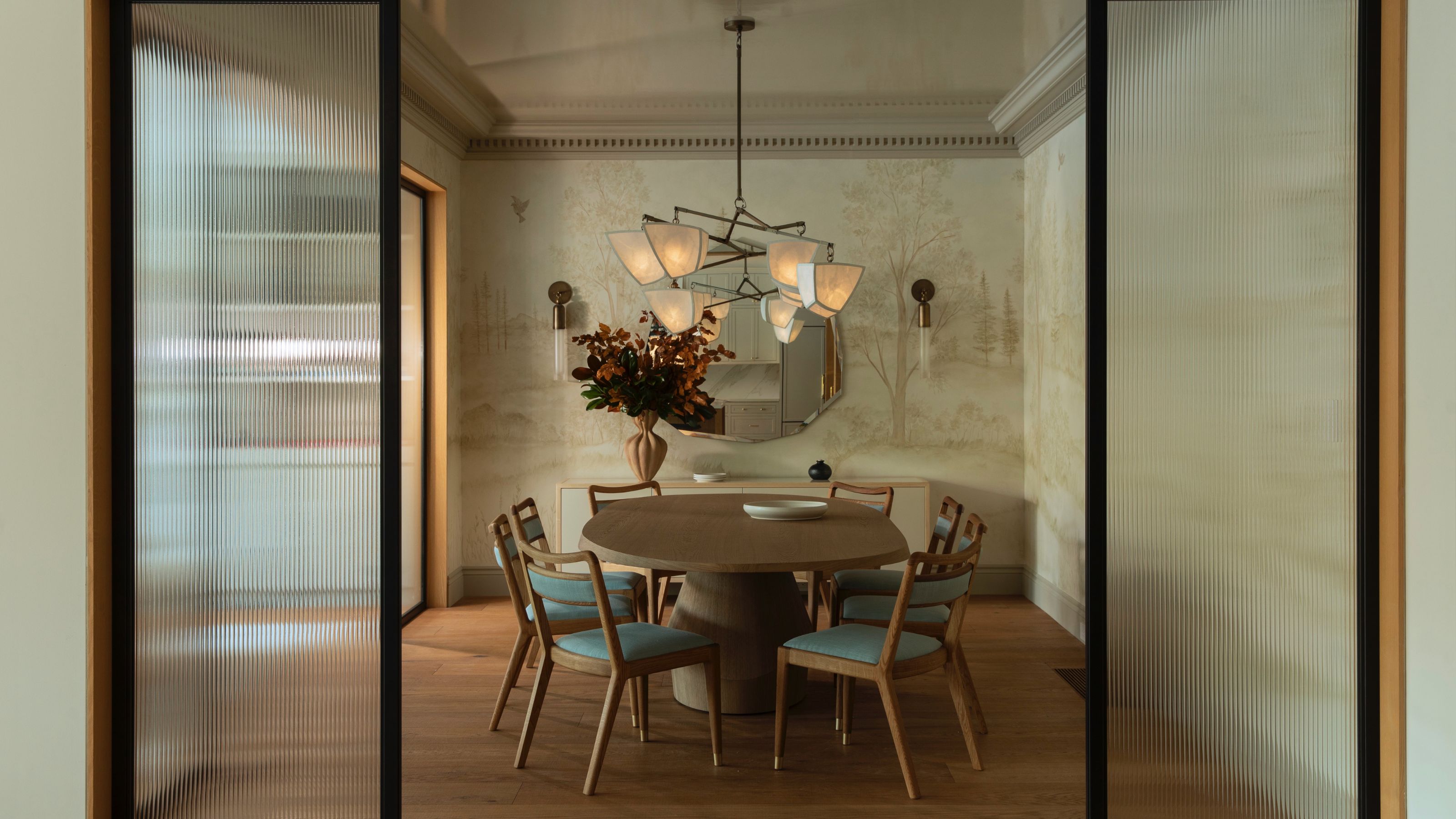 An elegant dining room seen through fluted glass sliding doors, featuring a round wooden pedestal table surrounded by mid-century modern chairs with light blue upholstery. The room is decorated with a scenic landscape mural wallpaper, a large sculptural chandelier with white shades, and a minimalist sideboard under a round mirror