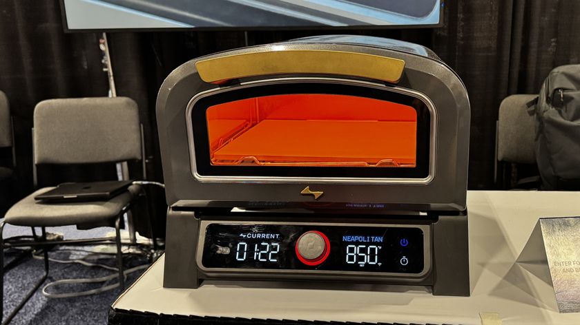 Current Model P pizza oven on a white table with orange light inside with chairs and black curtain in the background