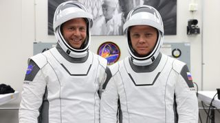 Two astronauts in sleek white and black spacesuits with helmet visors up.