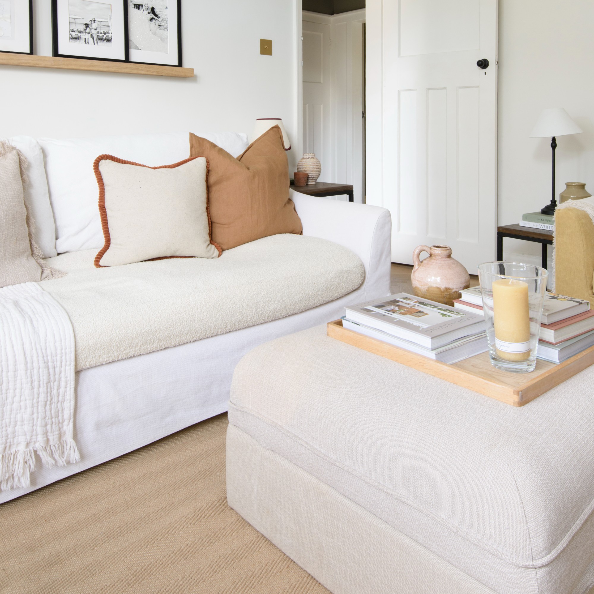 A white living room with a white sofa and a cream ottoman used as a coffee table with a wooden tray on top