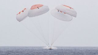 A space capsule makes an ocean splashdown under four orange and white parachutes