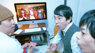 A screenshot from the movie "Beyond The Infinite Two Minutes" where three men look surprised around an apple computer