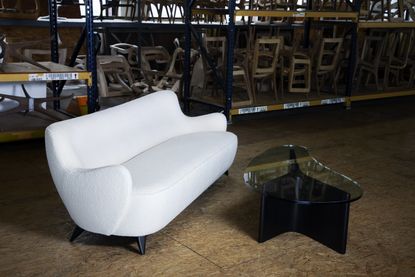 Vladimir Kagan furniture shown inside a warehouse space: 1950s white upholstered sofa with sinuous shape, and coffee table with Y-shaped wooden base and glass top