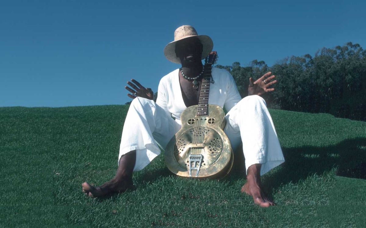 Taj Mahal sitting barefoot with guitar on a grass bank