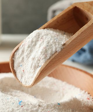 A close-up shot of a wooden scoop with white laundry powder being lifted from a wooden bowl