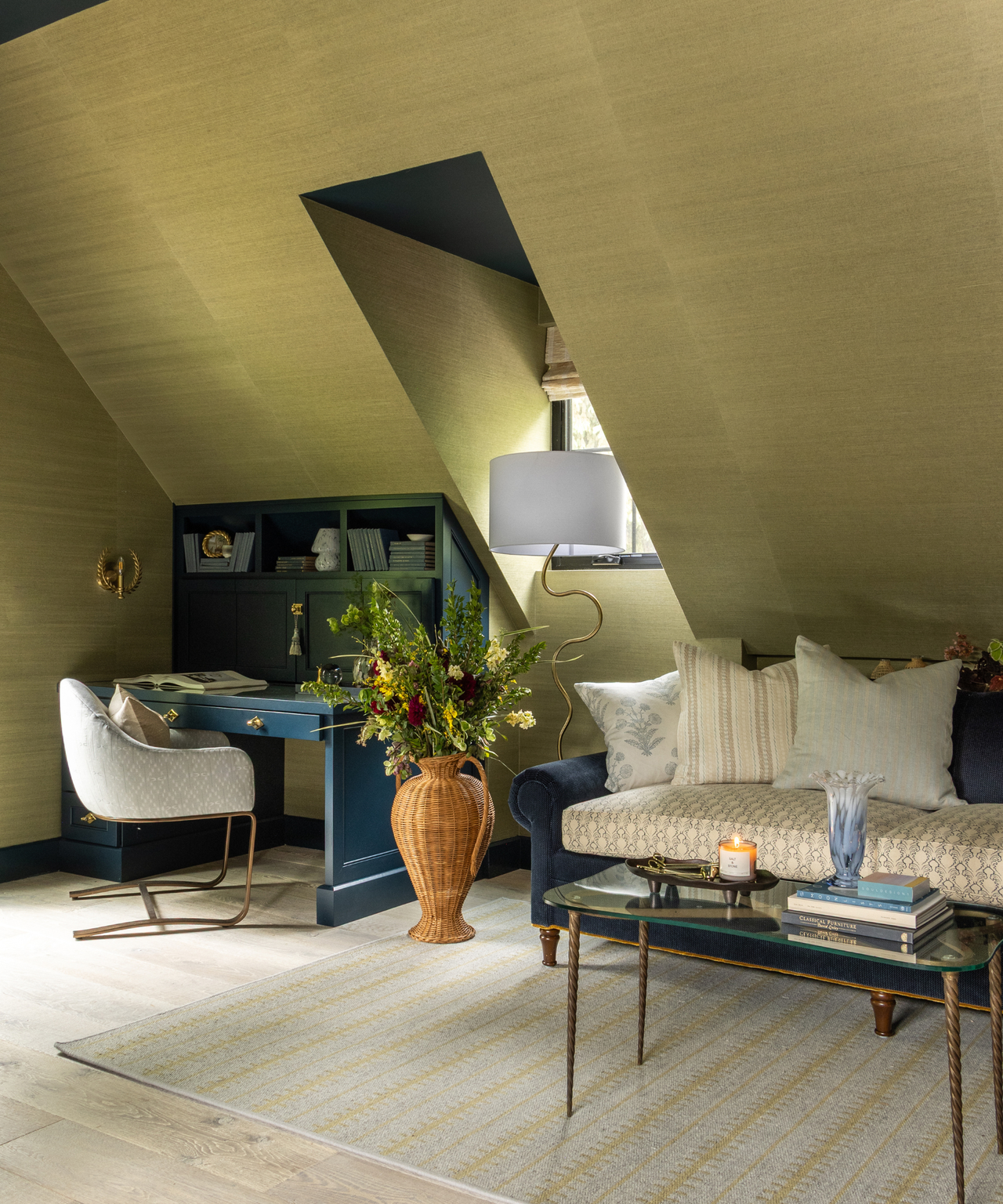 a room with gold grass cloth wallpaper, a couch, a coffee table, a vase with flowers, a built in desk, and a desk chair
