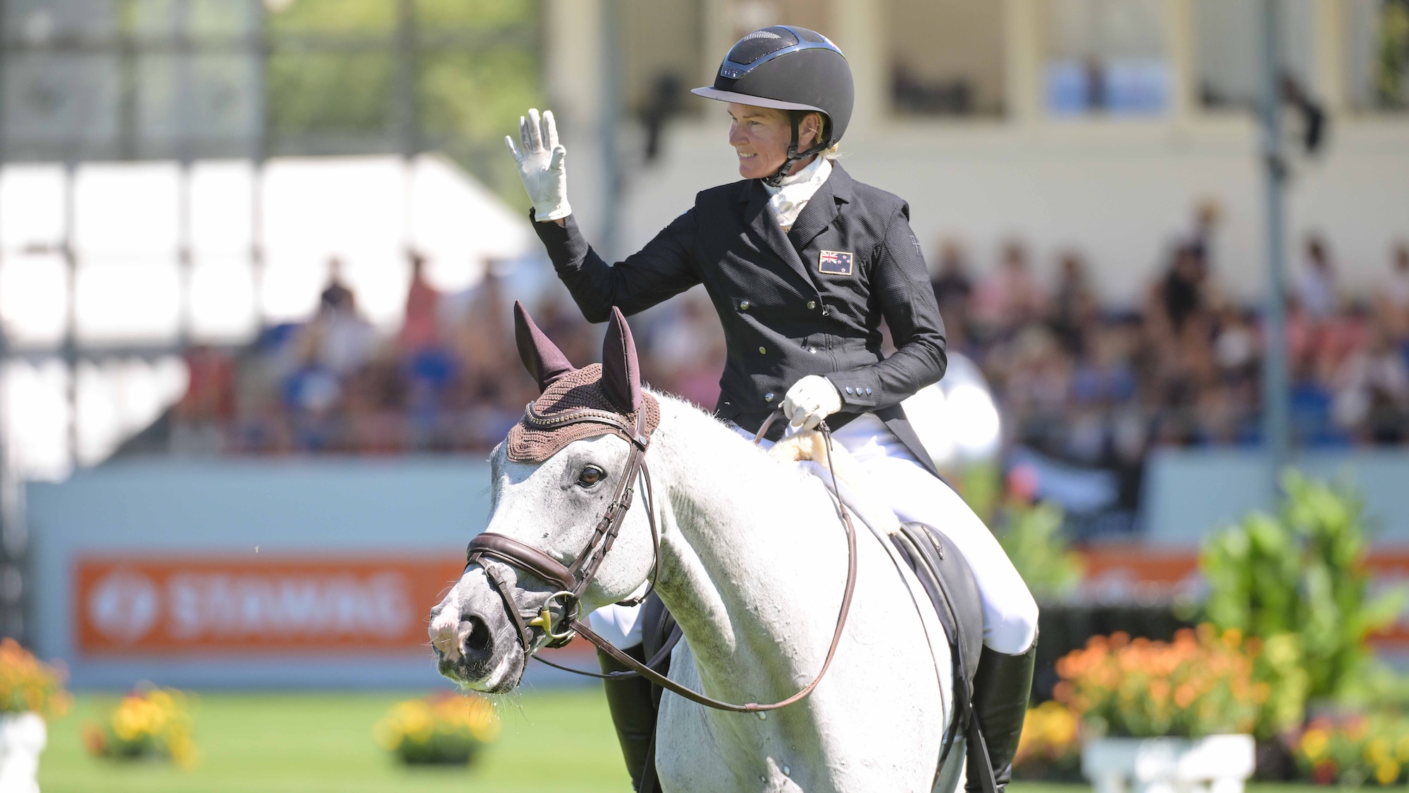 Image shows rider Jonelle Price waving to the crowds, as her grey horse Senor Crocodillo walks out of the arena on a long rein