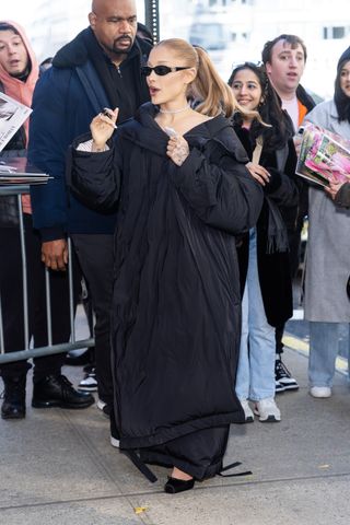 A photo of Ariana Grande pairing her sleeping bag coat with a diamond choker, black Gucci sunglasses, and black satin balletcore pumps.