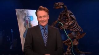 Conan O'Brien stands smiling in front of props in The Tonight Show With Conan O'Brien.