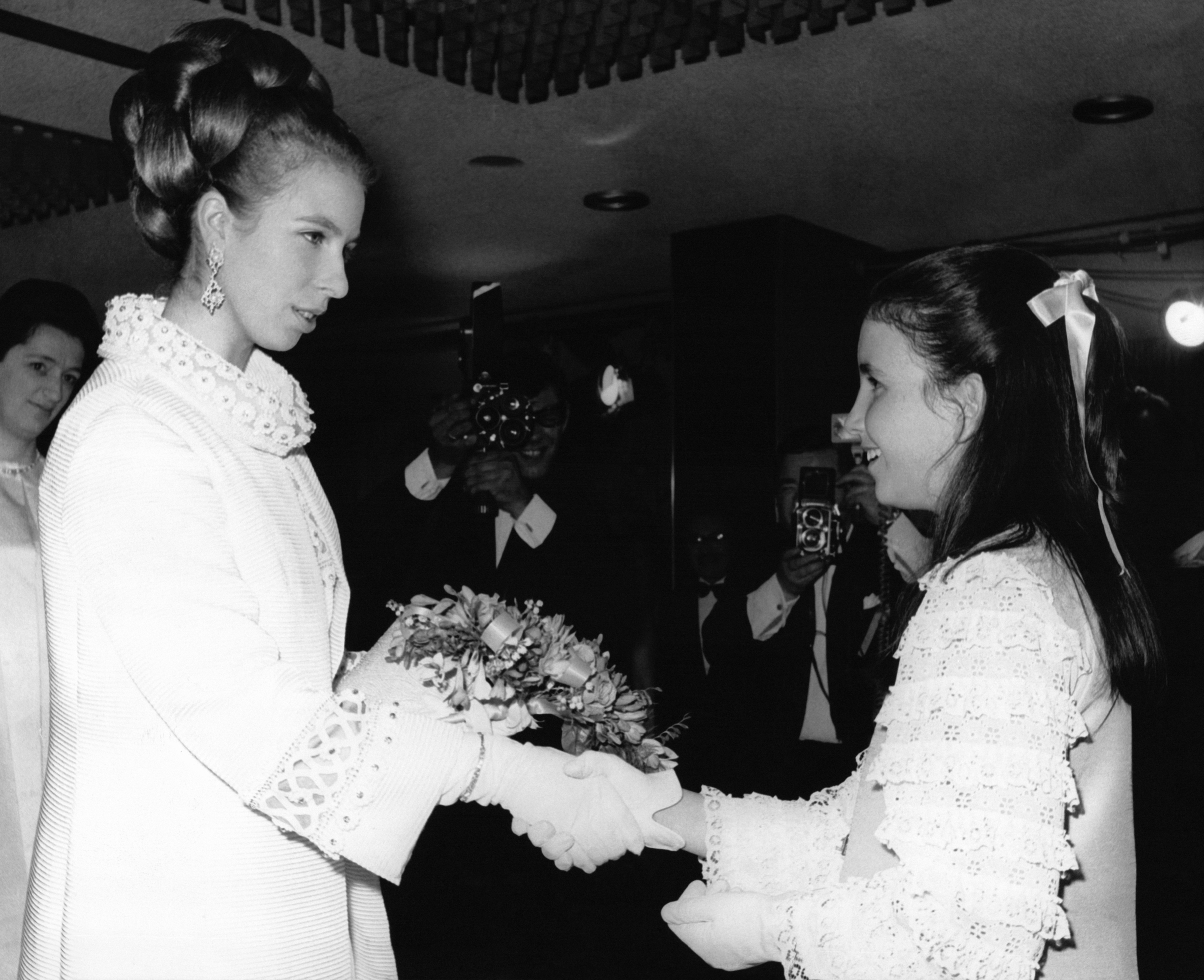 Princess Anne wearing a white coat shaking hands with a girl in 1969