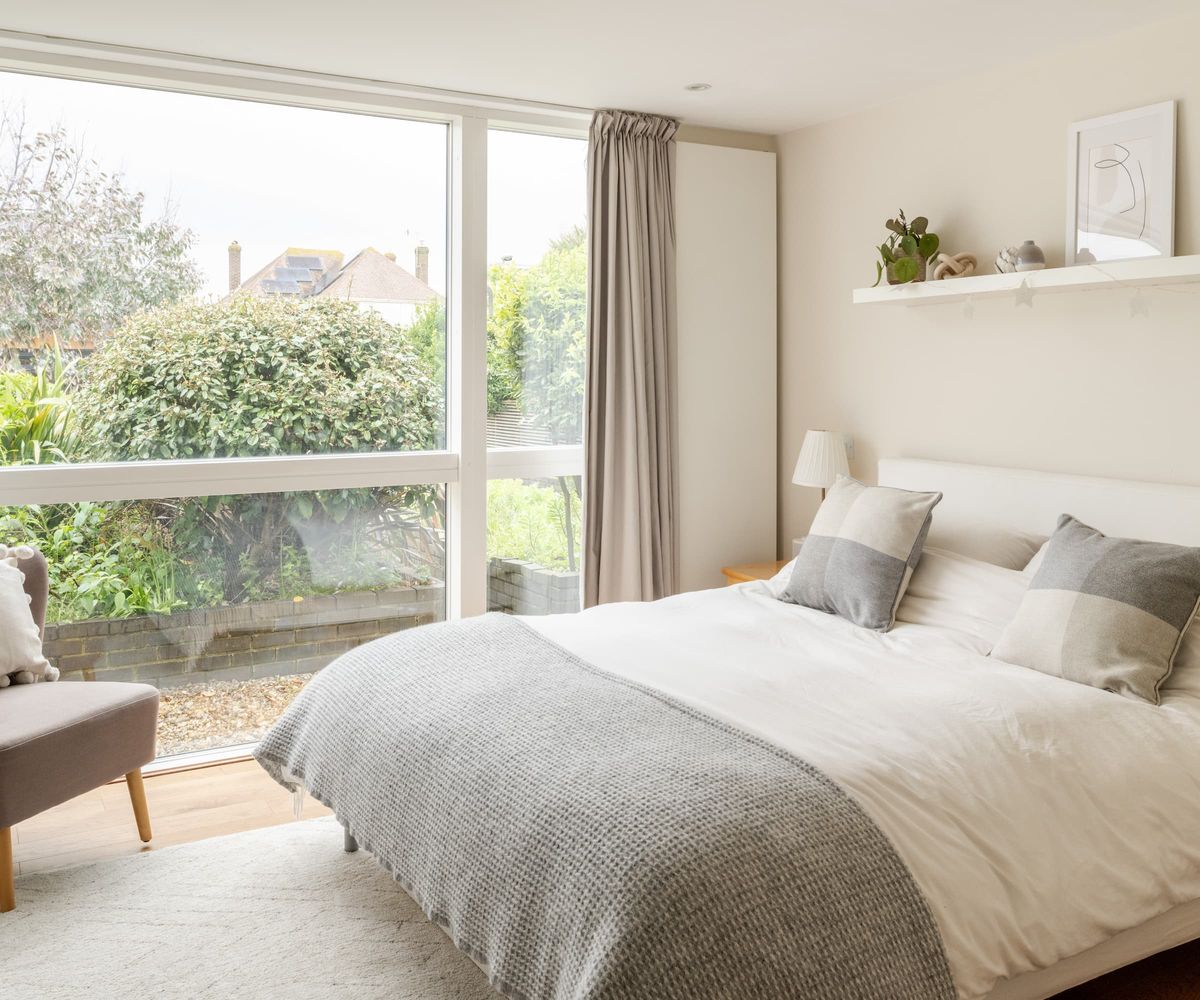 A white bedroom with a large picture windows looking onto a gravelled garden