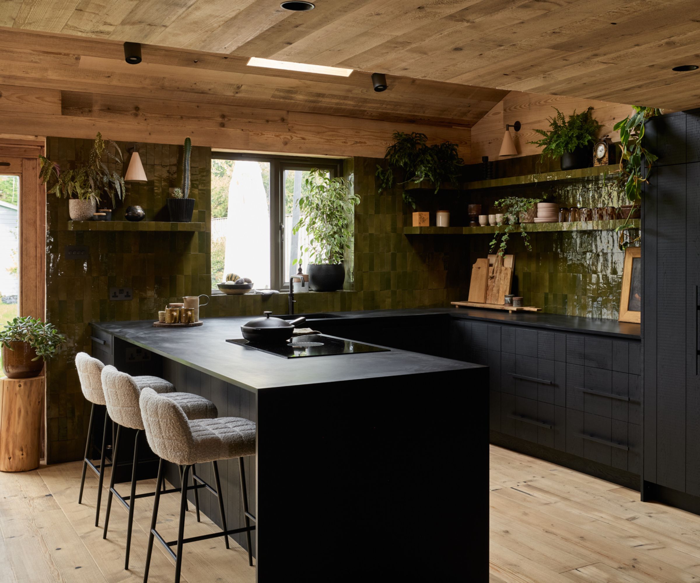 Dark kitchen with black island, green tiled walls and timber ceiling. Bar stools line the island, with plants and open shelving adding texture.