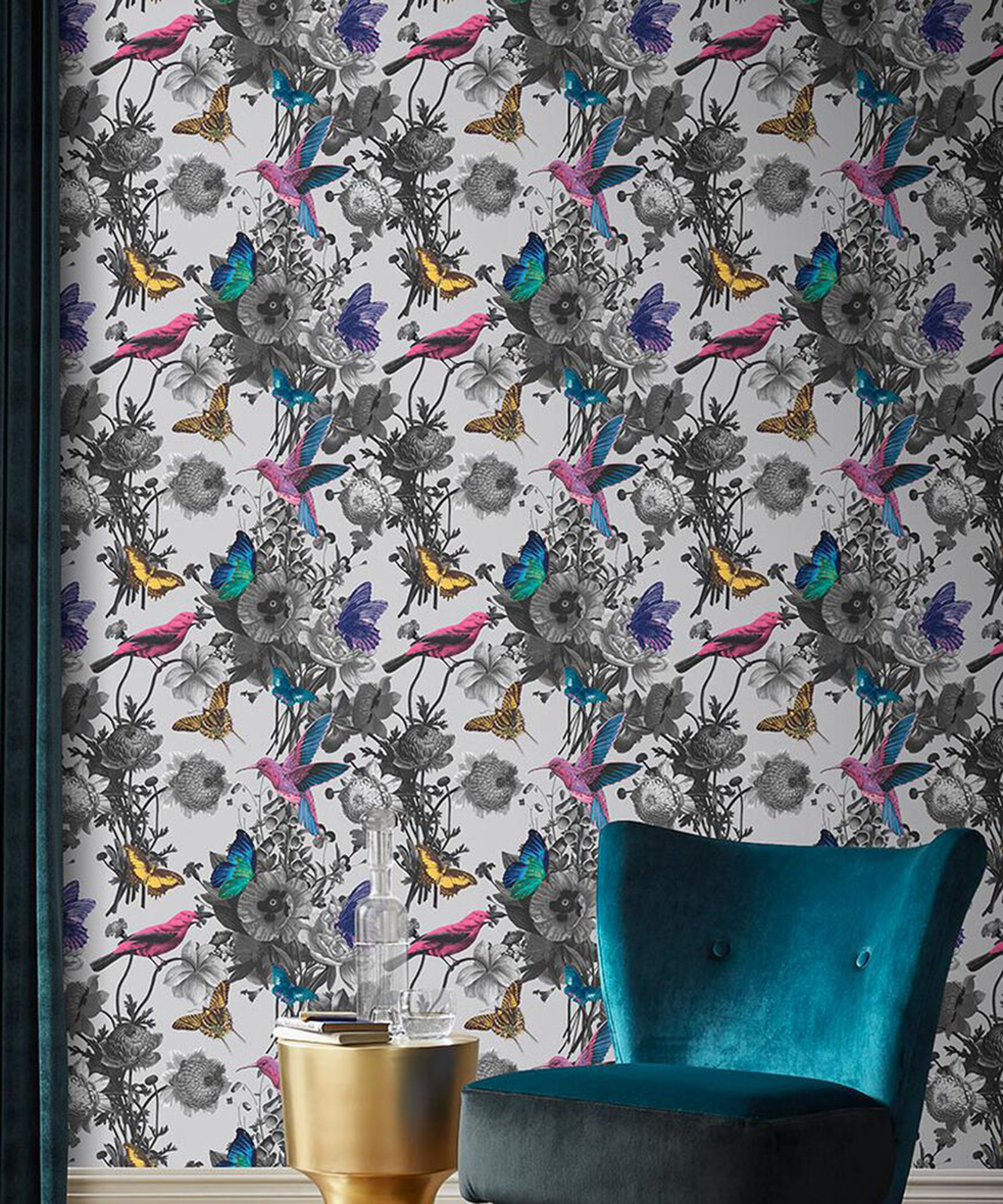 A grey patterned wallpaper by Graham & Brown with birds and floral design with teal velvet chair, velvet curtains and metallic gold side table with glass bottle filled with water