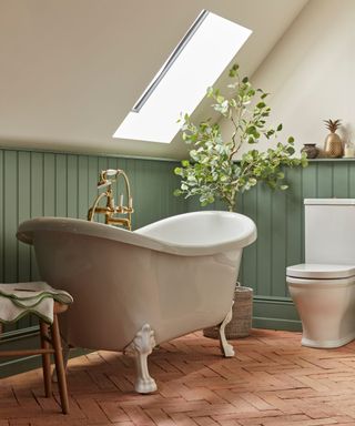 A charming bathroom with a classic freestanding clawfoot bath positioned under a slanted skylight. The space features sage green tongue-and-groove wall panelling and reclaimed terracotta floor tile.