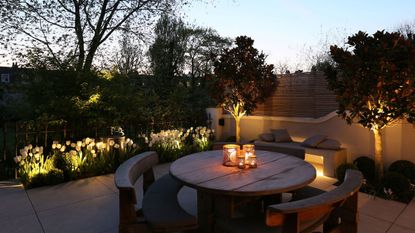 An outdoor area illuminated at dusk with a round dining table with a round curved bench around it and a built-in bench seat behind it against a white wall with trees and planting all lit up
