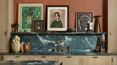 kitchen countertop and backsplash with green marble and a picture ledge with artworks and sculpture, brass taps, blonde timber cabinetry, and terracotta limewashed walls