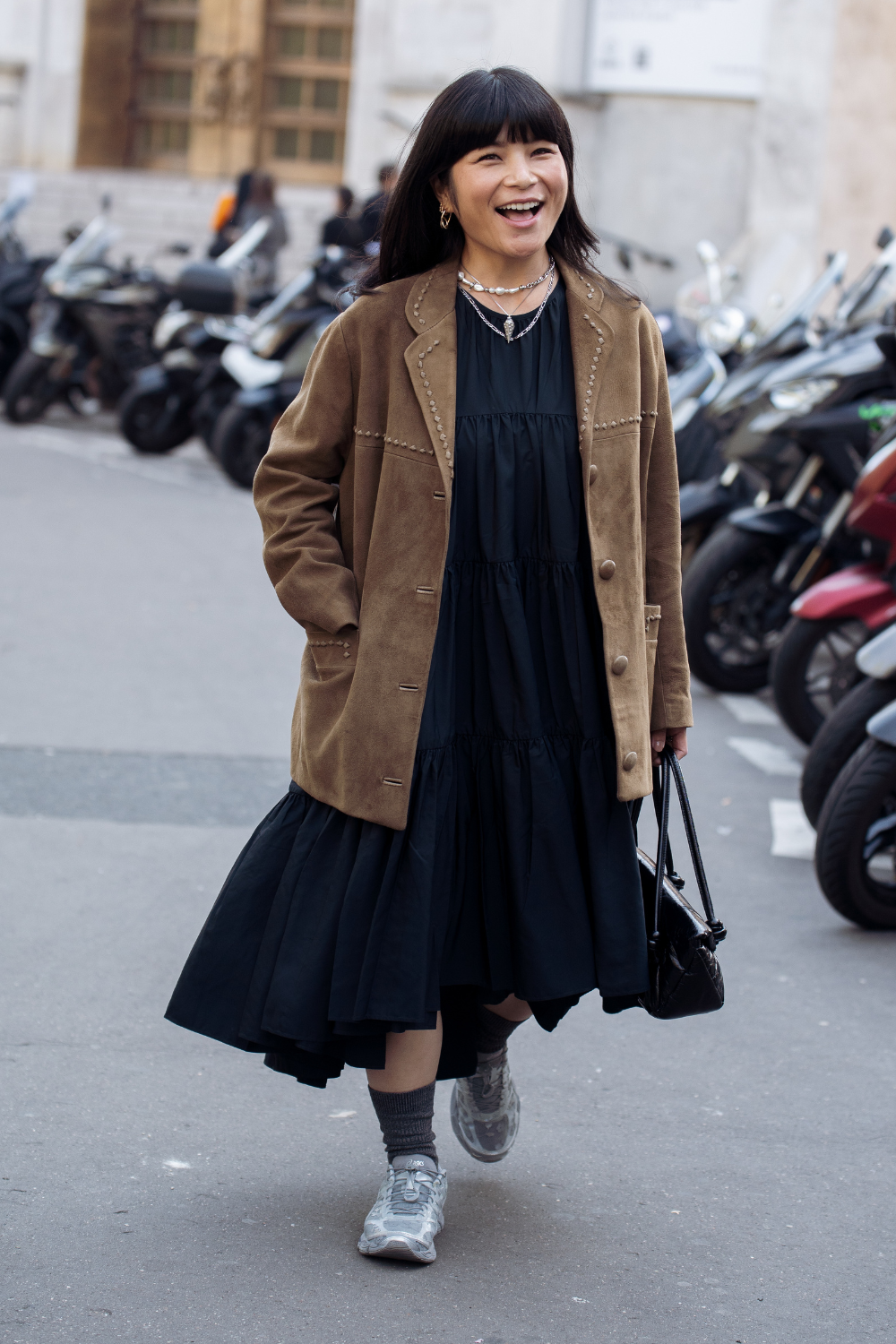 Elie Inoue wears a black tiered midi dress under a brown suede blazer with studded details, grey socks with silver sneakers, layered necklaces, and carries a black leather handbag outside Cecilie Bahnsen show during the Womenswear Spring Summer 2026 as part of Paris Fashion Week