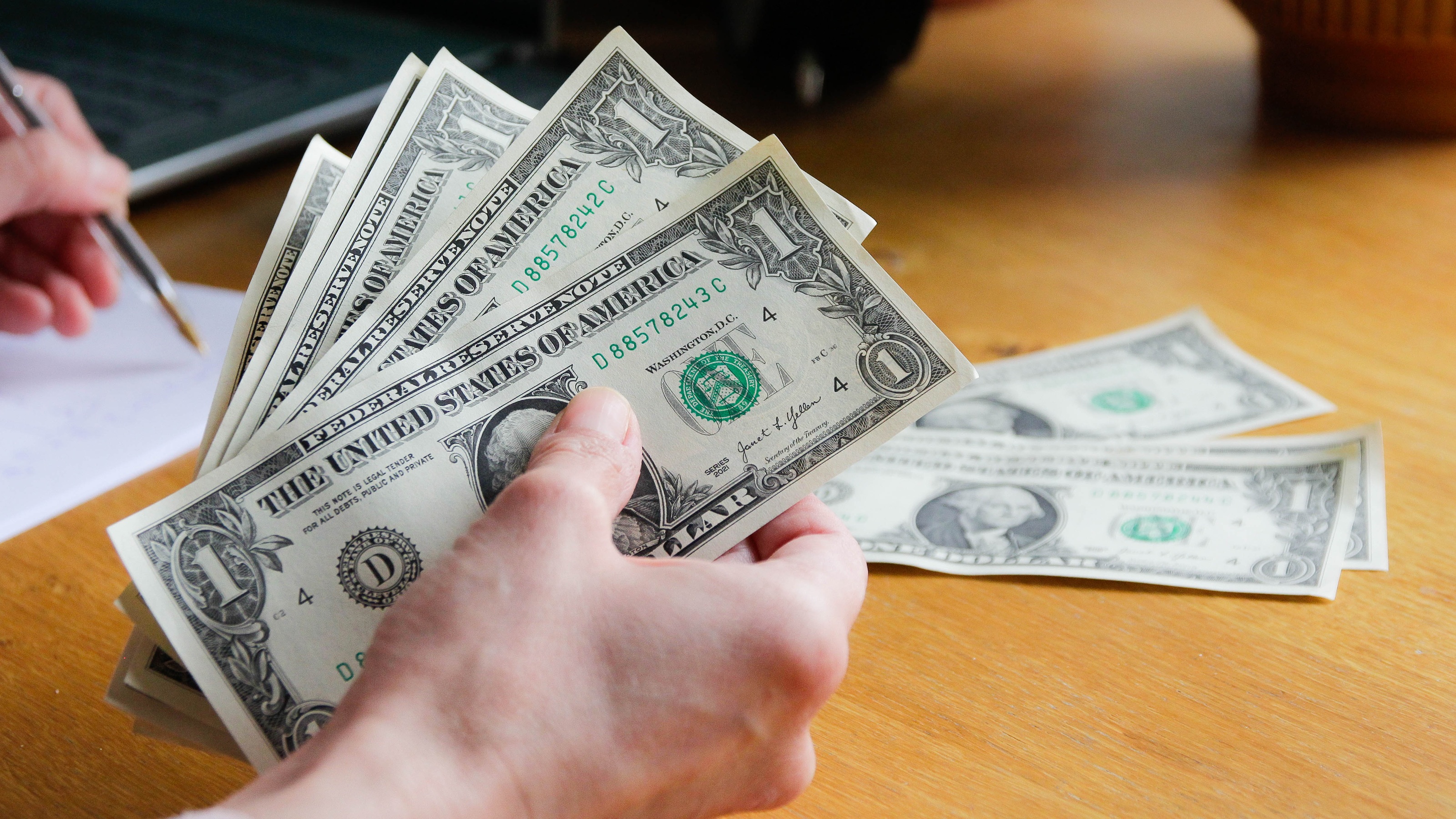 A man's hand holds several dollar bills while he makes notes on a piece of paper.