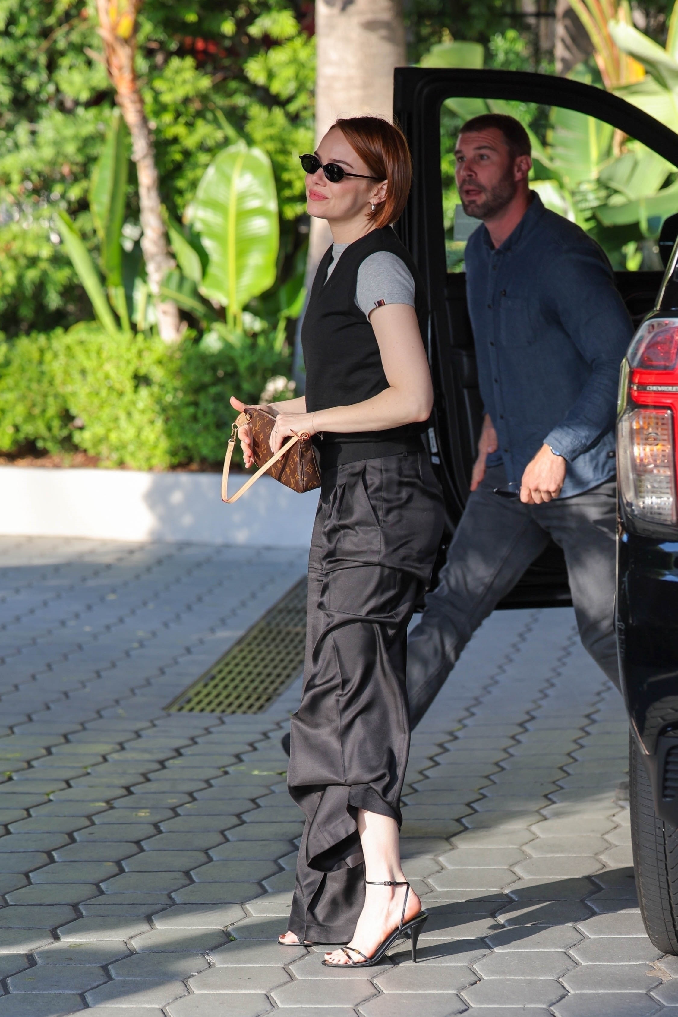 Emma Stone wears black parachute pants, black strappy heels, a black preppy vest, a gray t-shirt, and carries a Louis Vuitton bag