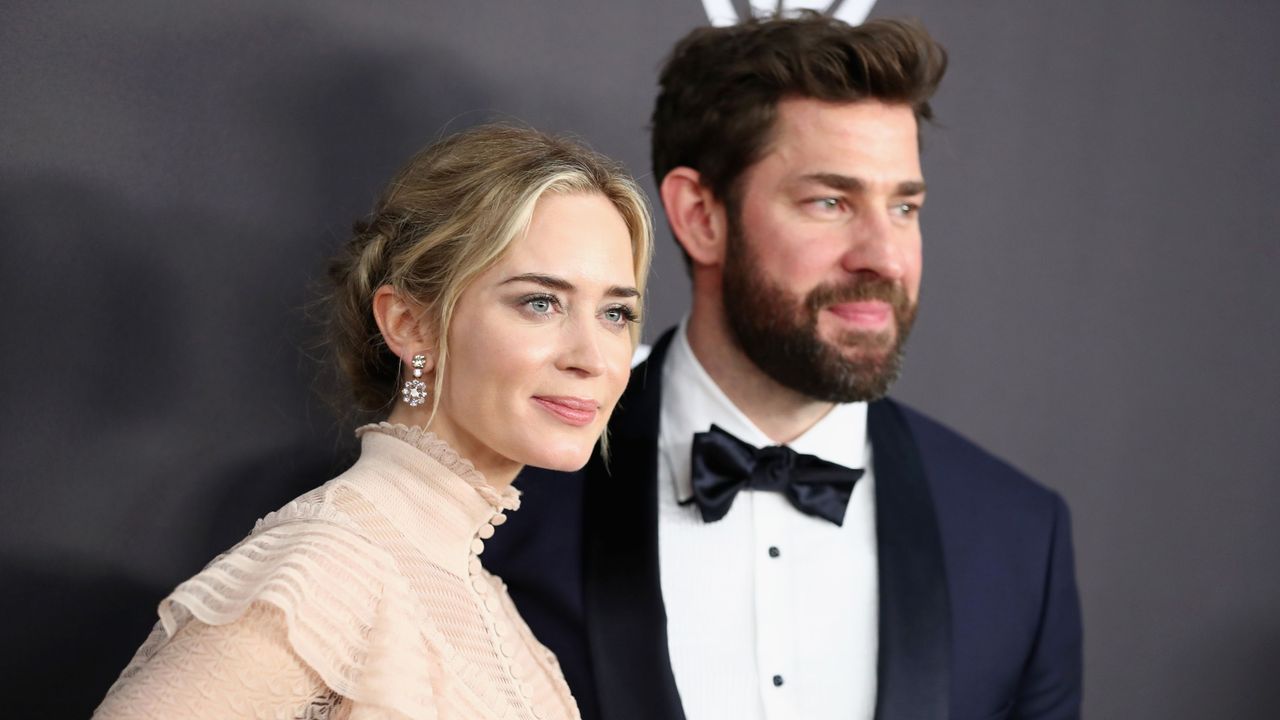 Emily Blunt wearing a beige dress with a blonde up-do and John Krasinski wearing a black suit, bow-tie, and white shirt, in front of a black background