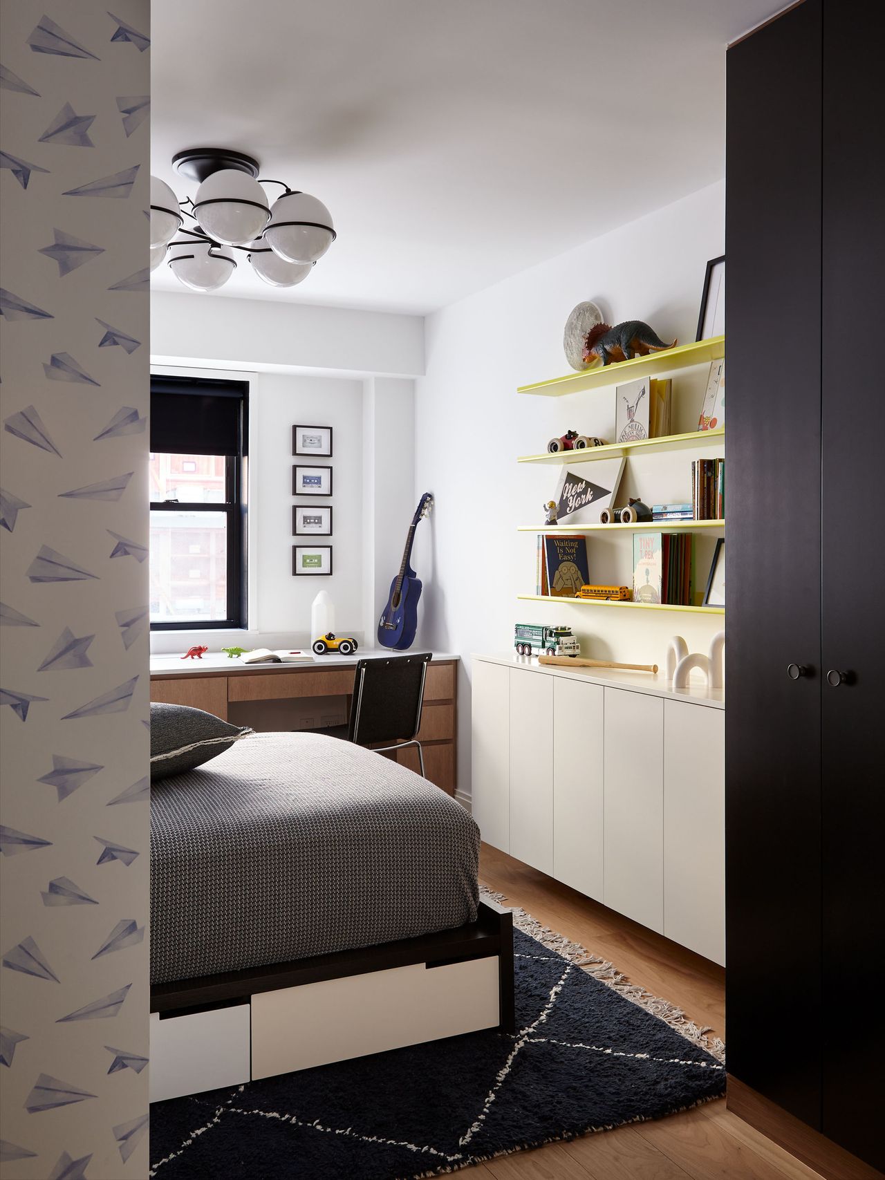 a boys bedroom with yellow floating shelves, a black storage unit and wallpaper on the wall in the foreground with dark gray bedding on the bed and a blue guitar sitting atop the desk in the background
