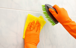 Rubber gloved hands cleaning shower tiles with brush and yellow cloth