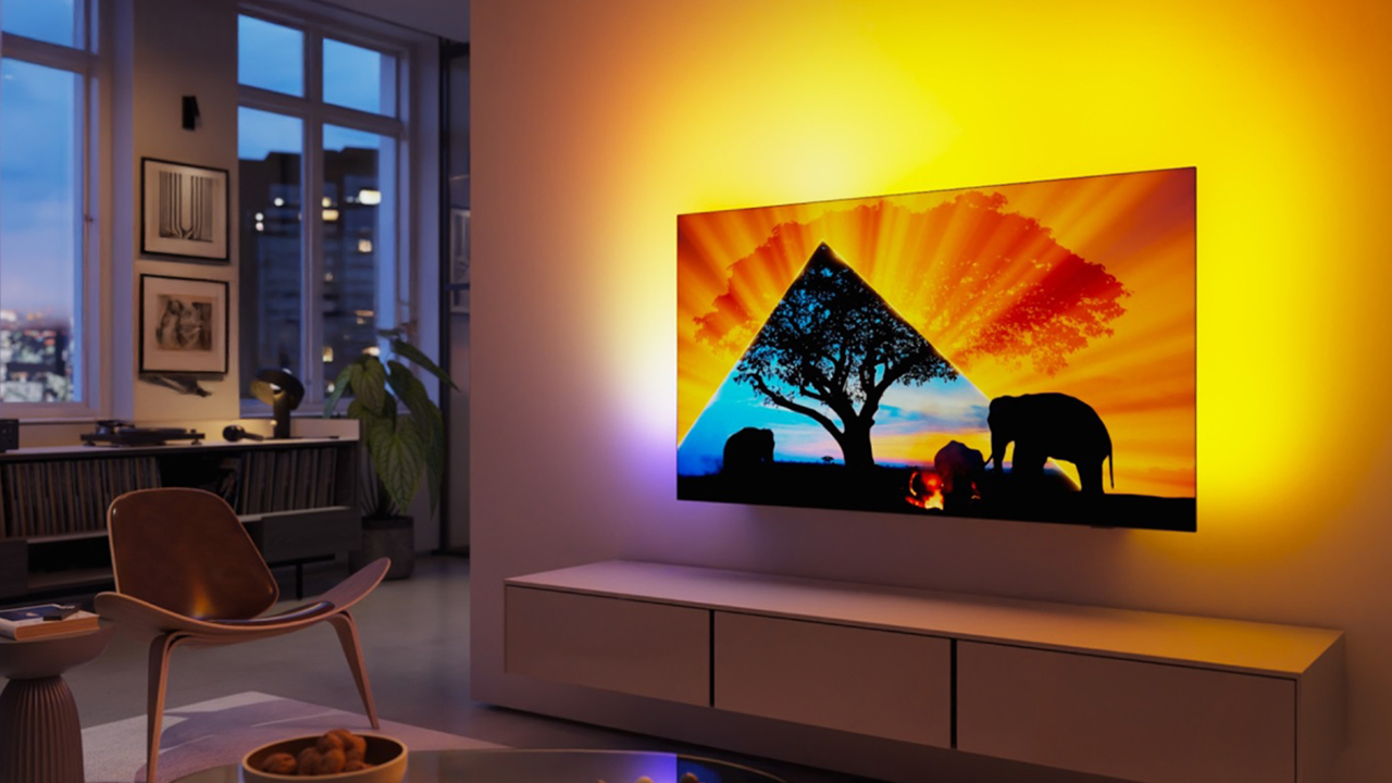 Philips OLED 759 wall-mounted with Ambilight scattering yellow and blue light onto the wall behind, and a living room scene to the left