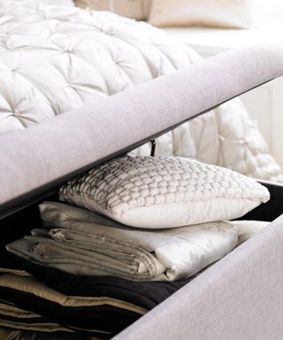 A gray storage ottoman next to a white bed with pillows and sheets inside it