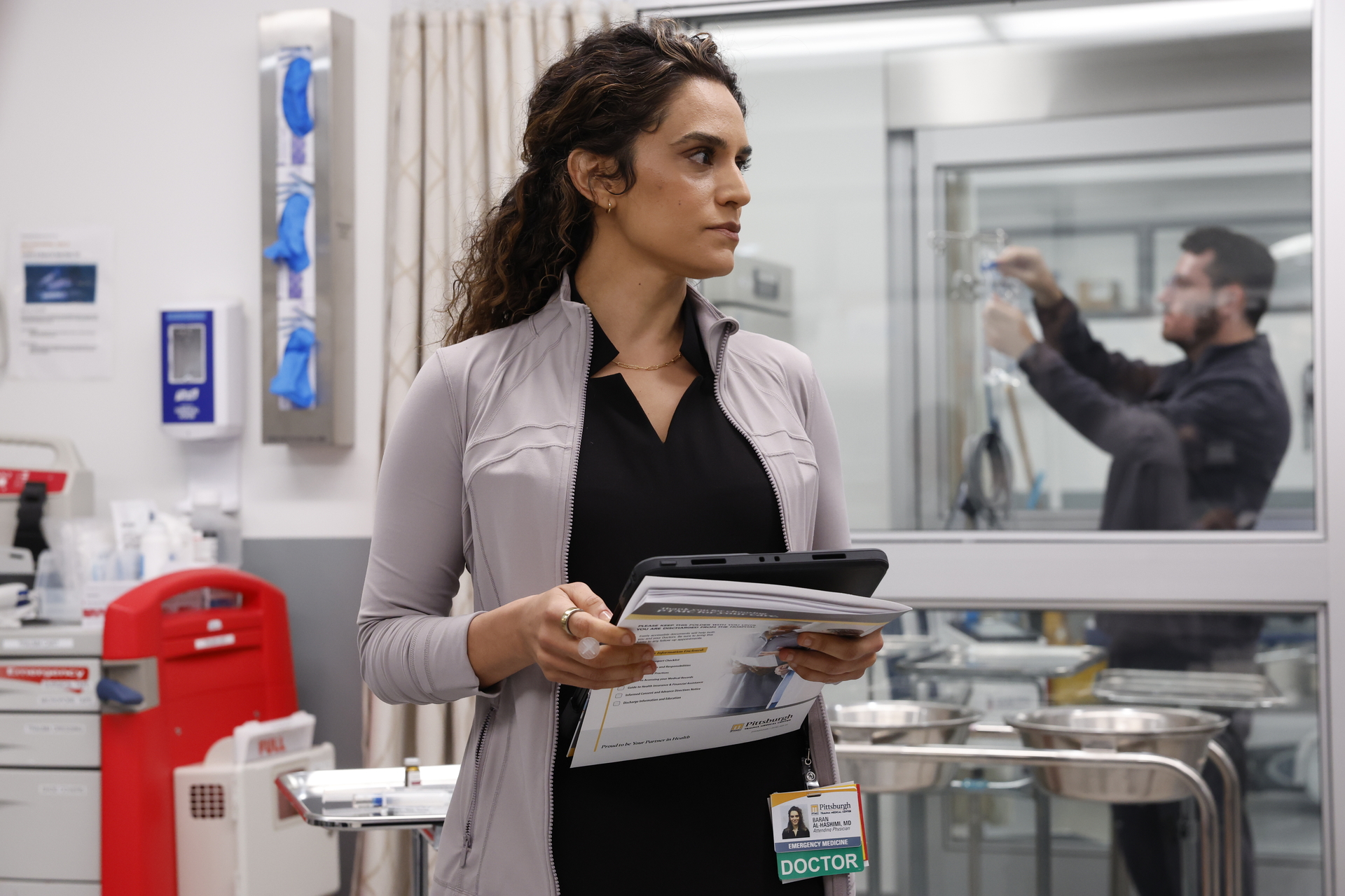 sepideh moafi as dr. al-hasmini wearing a lavendar lulu lemon jacket over her black scrubs holding paper work in a doctor's office in the pitt season 2