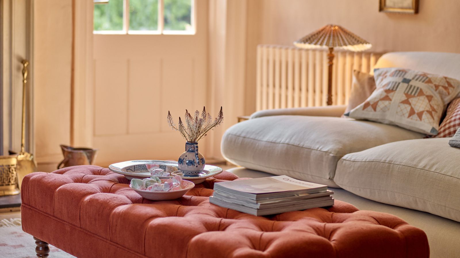 A red upholstered ottoman in a warm living room, with a beige sofa behind it. A small floor lap with a pleated shade beside it. Some magazines and trays on top of the ottoman. 