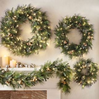 Faux Lit Mixed Greens Wreath & Garland With Bells on a white mantel.