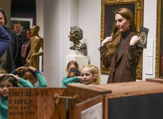 Kate Middleton wearing a brown blazer putting her hands on her shoulders and holding a pamphlet while playing a game with a group of kids in teal sweatshirts at the National Portrait Gallery in London