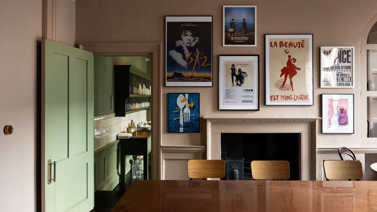 A dining room with gallery wall showing vintage posters in frames on plaster pink walls above and around fireplace, view into kitchen which is apple green, large dining table with three vintage style school chairs