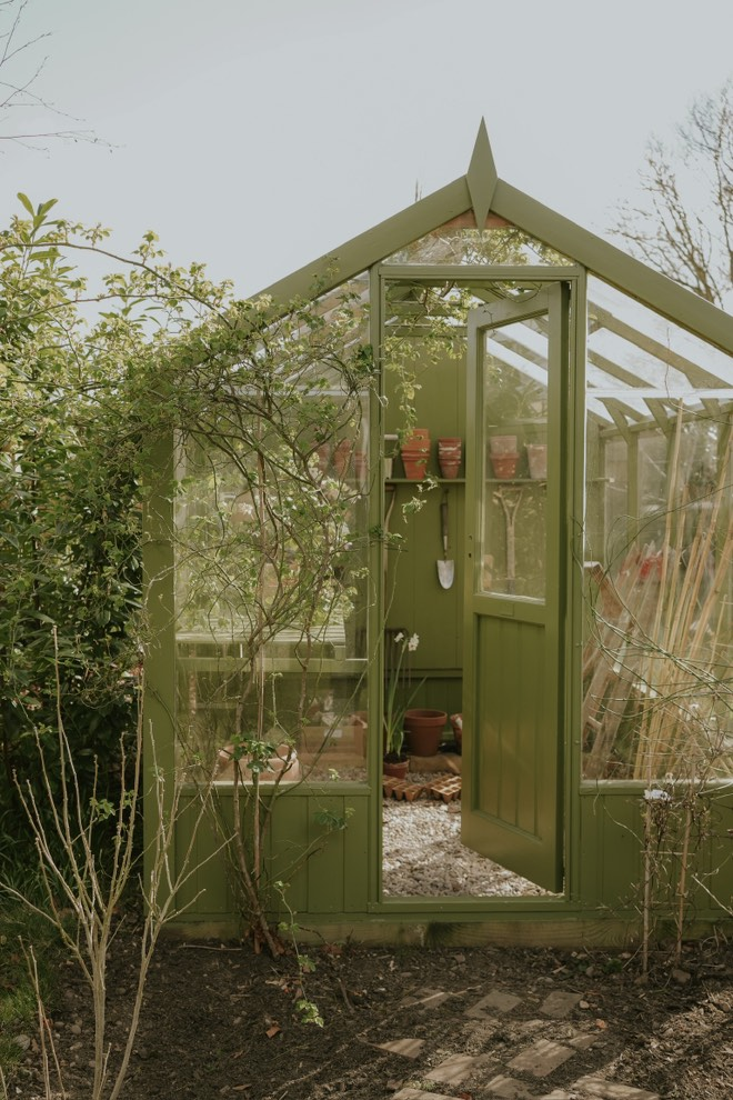 Image of a wooden and glass greenhouse in a garden. The structure is painted a bright olive green and there are terracotta pots on the shelves inside.