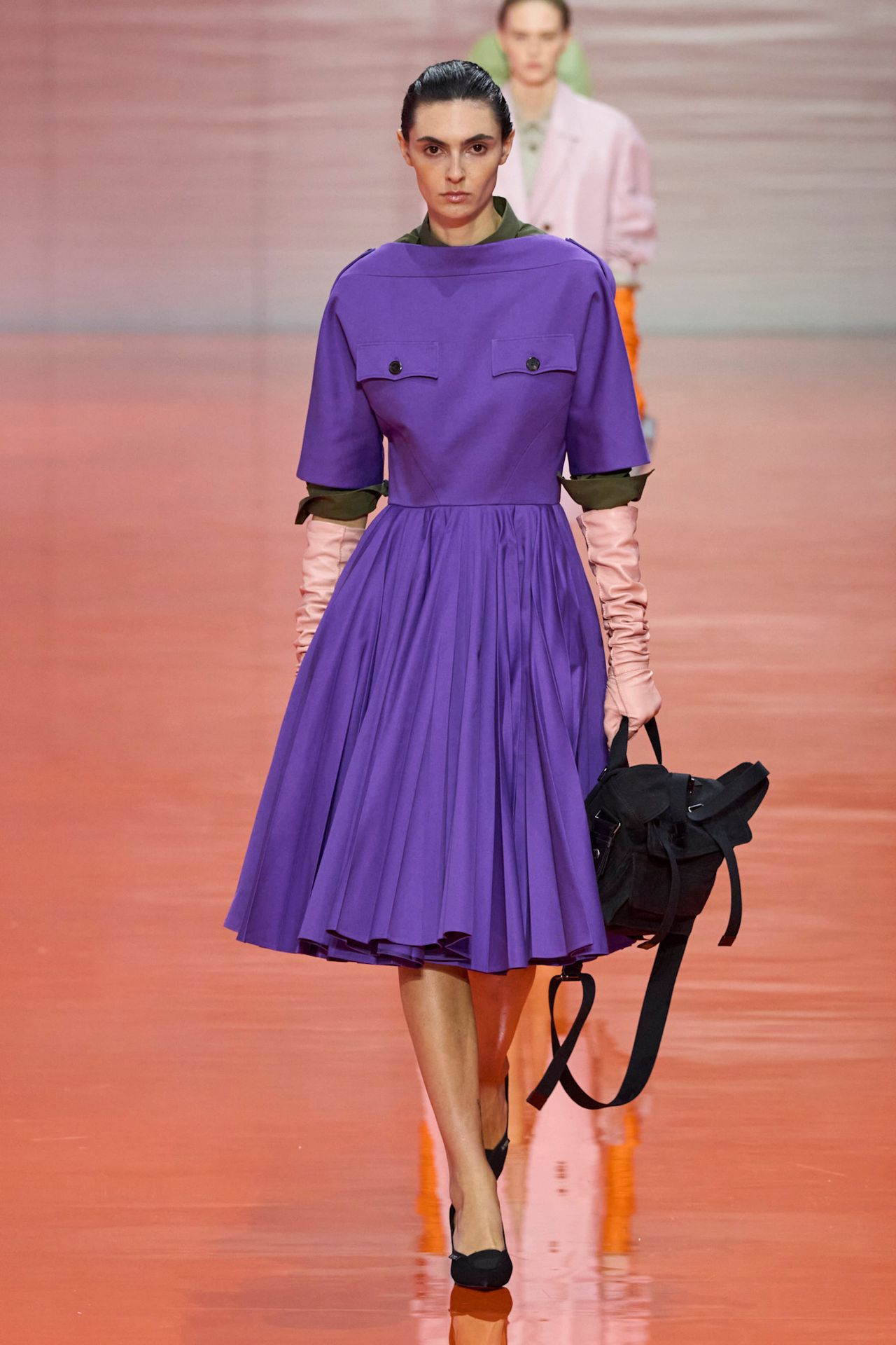 A Prada model wearing a purple boat-neck dress with a voluminous pleated skirt, an olive green shirt underneath, pale pink leather opera gloves, a black nylon backpack, and black pointed kitten heels.