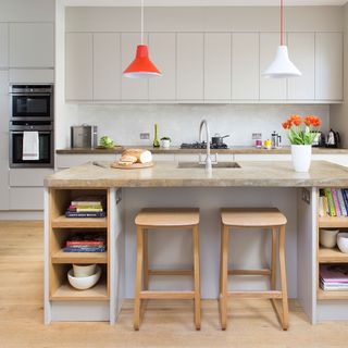 stone coloured island with wooden stools and flooring, red and white pendants and light grey cabinets