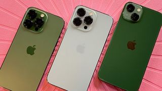 Alpine Green iPhone 13 Pro, Sierra Blue iPhone 13 Pro, green iPhone 13 face down on pink placemat with camera bumps facing up
