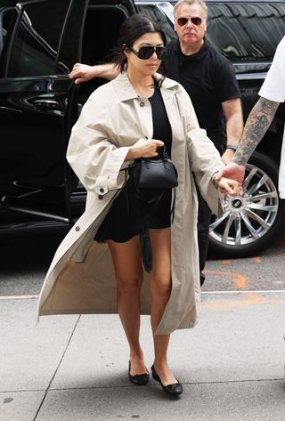 Kourtney Kardashian wears a tan trench coat with a black short skirt and black ballet flats