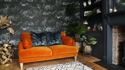 A living room with a wall covered in a dark floral wallpaper with an orange velvet sofa in front of it