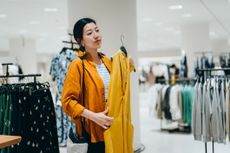 Young Asian woman shopping in a clothing store, looking at a yellow dress in mirror.