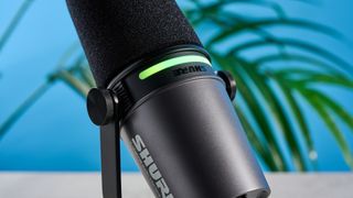 the shure mv7i, a black microphone with a black muff, touch LED pad, XLR connection and TRS input and USB-C input at the rear, photograhped against a blue tom's guide background with a pair of over-ear wired headphones