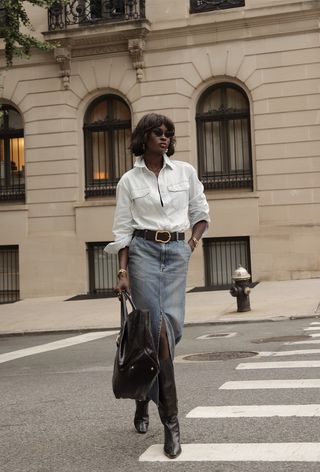 A denim skirt outfit is shown in a photo of a woman on the street wearing a light wash denim skirt with a black belt, denim midi skirt, black knee-high boots, and a black handbag