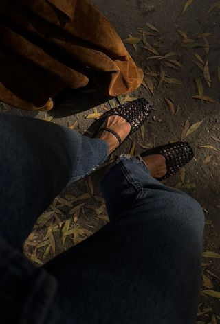 The woven leather shoe trend is shown in a close photo of a woman feet where she's standing outside wearing skinny jeans, a suede jacket, and black woven ballet flats