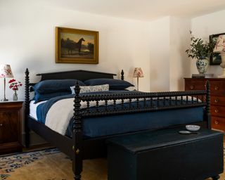 bedroom with black spool bed, navy and white bedlinen, dark blue trunk, wooden chest of drawers, horse painting and neutral rug